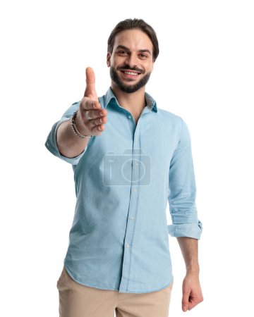 Foto de Feliz joven con barba en camisa de mezclilla apuntando de la mano hacia adelante y saludando mientras sonríe y posa sobre fondo blanco - Imagen libre de derechos