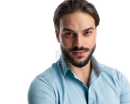 Foto de Retrato de joven guapo con barba vistiendo camisa de mezclilla azul, mirando hacia adelante y posando delante de fondo blanco - Imagen libre de derechos