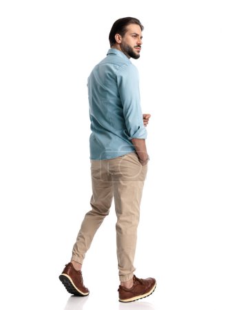 Foto de Vista trasera de un joven casual caminando con la mano en el bolsillo y mirando hacia un lado delante del fondo blanco - Imagen libre de derechos