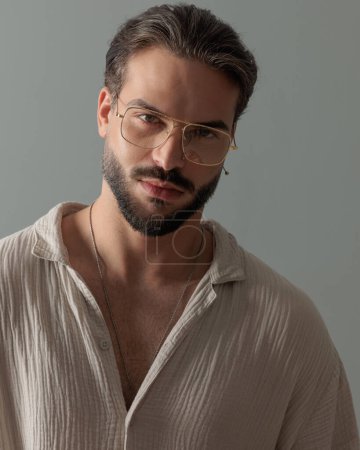 Téléchargez les photos : Portrait d'un bel homme aux lunettes tournées vers l'avant et posant devant un fond gris - en image libre de droit