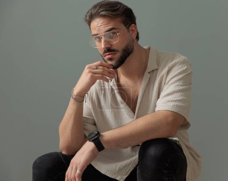Téléchargez les photos : Cool barbu gars avec des lunettes tenant les coudes sur les genoux et regardant vers l'avant tout en pensant en face de fond gris - en image libre de droit