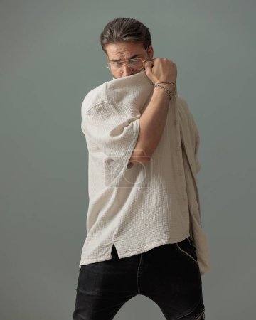 Photo for Cool young man with glasses covering face with shirt and posing in front of grey background - Royalty Free Image