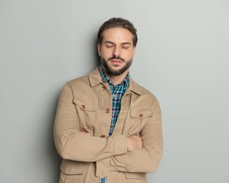 Photo for Tired young man in beige jacket closing eyes and resting while crossing arms and posing in front of grey background - Royalty Free Image