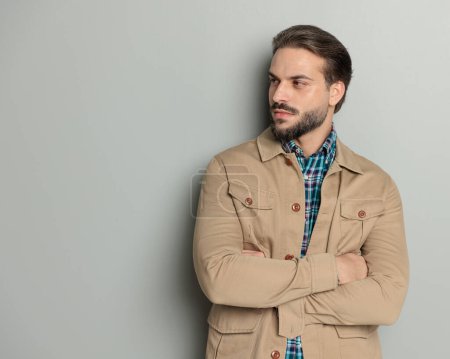 Foto de Joven confiado en chaqueta beige cruzando brazos y mirando a un lado mientras posa frente a fondo gris - Imagen libre de derechos