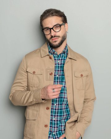 Foto de Sexy joven barbudo hombre en chaqueta beige con gafas mirando hacia otro lado mientras sostiene la mano en el bolsillo sobre fondo gris - Imagen libre de derechos