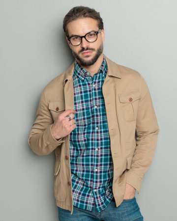 Foto de Sexy hombre de moda con gafas sosteniendo y ajustando chaqueta beige y sosteniendo la mano en el bolsillo sobre fondo gris - Imagen libre de derechos