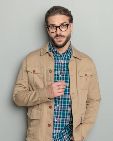 Foto de Sexy barbudo casual hombre en chaqueta beige cogido de la mano en los bolsillos y posando delante de fondo gris - Imagen libre de derechos