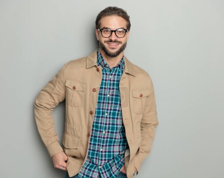 Téléchargez les photos : Heureux homme décontracté avec des lunettes tenant la main dans les poches et souriant devant fond gris - en image libre de droit