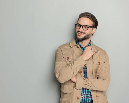 Photo for Happy casual guy with glasses in beige jacket folding arms and looking to side while smiling in front of grey background - Royalty Free Image
