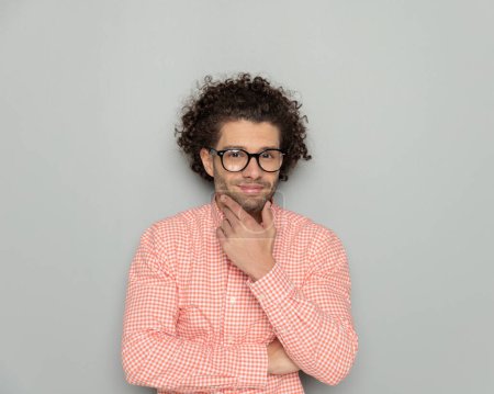 Téléchargez les photos : Portrait d'un homme confiant avec des lunettes portant une chemise à carreaux touchant le menton et se demandant en posant devant un fond gris - en image libre de droit