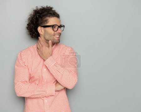 Photo for Attractive curly hair guy with glasses folding arms and looking to side while touching neck in front of grey background - Royalty Free Image
