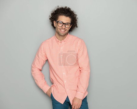 Foto de Retrato de hombre casual feliz con gafas cogidas de la mano en los bolsillos y sonriendo mientras posando frente a fondo gris - Imagen libre de derechos