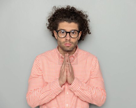 Téléchargez les photos : Portrait de jeune homme sérieux avec des lunettes regardant vers l'avant et priant pour un miracle devant fond gris - en image libre de droit