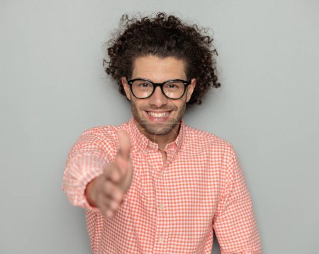 Téléchargez les photos : Heureux bouclé cheveux gars avec des lunettes souriant tout en serrant la main devant fond gris en studio - en image libre de droit