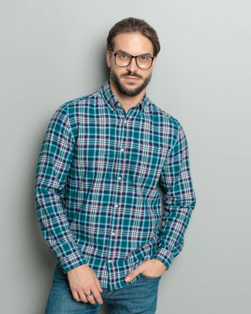 Foto de Atractivo joven con camisa a cuadros con gafas cogidas de la mano en el bolsillo y mirando hacia adelante mientras posando sobre fondo gris - Imagen libre de derechos