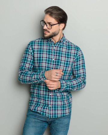 Foto de Joven guapo en camisa a cuadros ajustando las mangas mientras mira a un lado delante de fondo gris - Imagen libre de derechos