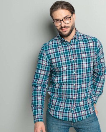 Foto de Hombre casual feliz con gafas con camisa a cuadros, sosteniendo la mano en el bolsillo trasero y sonriendo delante de fondo gris - Imagen libre de derechos
