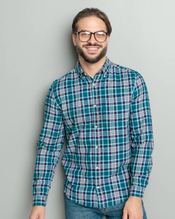 Foto de Joven feliz con camisa a cuadros y gafas y sonriendo delante de fondo gris en el estudio - Imagen libre de derechos