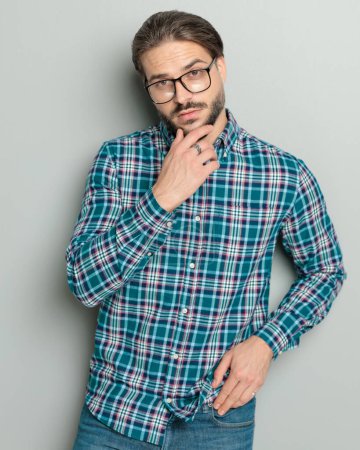 Foto de Hombre casual reflexivo en camisa con gafas tocando la barbilla y pensando mientras está de pie y posando delante de fondo gris - Imagen libre de derechos