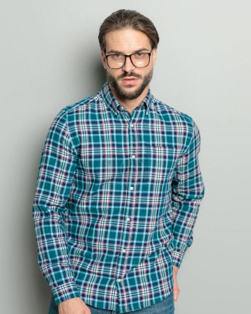 Foto de Sexy hombre sin afeitar con gafas en camisa a cuadros sosteniendo la mano en el bolsillo trasero y posando de una manera segura delante de fondo gris - Imagen libre de derechos