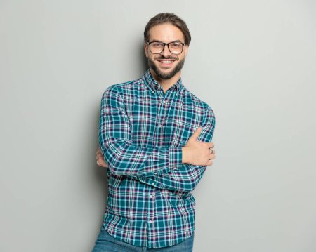 Foto de Chico casual excitado con gafas cruzando los brazos, siendo feliz y sonriente mientras posando frente a fondo gris - Imagen libre de derechos