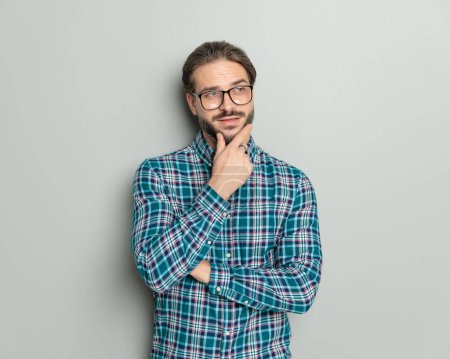 Foto de Retrato de hombre casual pensativo en camisa a cuadros con brazos plegables gafas y pensando en frente de fondo gris - Imagen libre de derechos