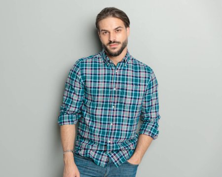 Foto de Confiado hombre casual con camisa a cuadros y la mano en los bolsillos, posando sobre fondo gris - Imagen libre de derechos