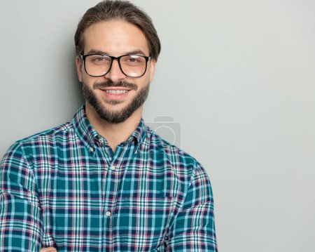 Foto de Retrato de hombre barbudo feliz en camisa a cuadros con gafas cruzando brazos y sonriendo delante de fondo gris - Imagen libre de derechos