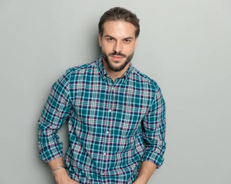 Foto de Retrato de atractivo hombre casual con camisa a cuadros, mirando hacia adelante y posando de una manera segura sobre fondo gris - Imagen libre de derechos