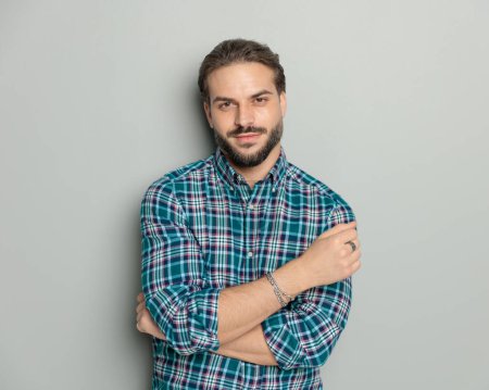 Foto de Retrato de joven confiado en camisa casual sonriendo mientras cruza los brazos y posando frente a fondo gris - Imagen libre de derechos