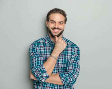 Foto de Hombre casual feliz en brazos plegables camisa a cuadros, tocando la barbilla y soñando mientras sonríe delante de fondo gris - Imagen libre de derechos