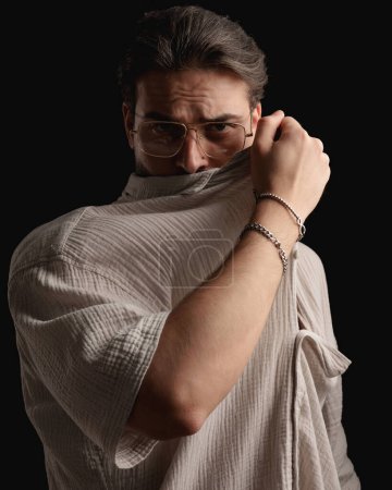 Photo for Sexy bearded man with glasses covering face with shirt and posing in front of black background - Royalty Free Image