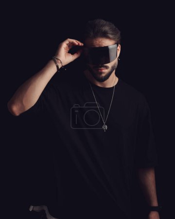 Téléchargez les photos : Beau jeune homme en t-shirt noir arrangeant cool lunettes de soleil de mode devant fond noir - en image libre de droit