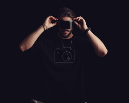 Photo for Cool young man in black t-shirt looking forward and adjusting sunglasses in front of black background - Royalty Free Image