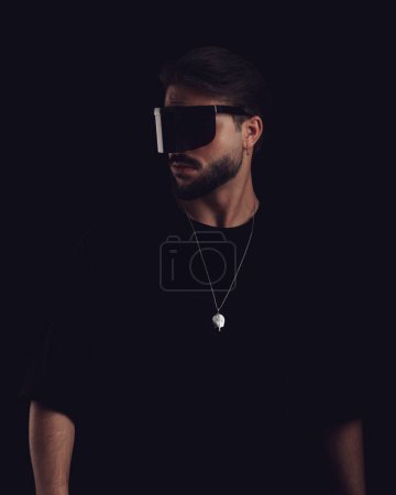 Téléchargez les photos : Jeune homme sexy en t-shirt portant des lunettes de soleil cool et regardant vers le côté en face de fond noir - en image libre de droit