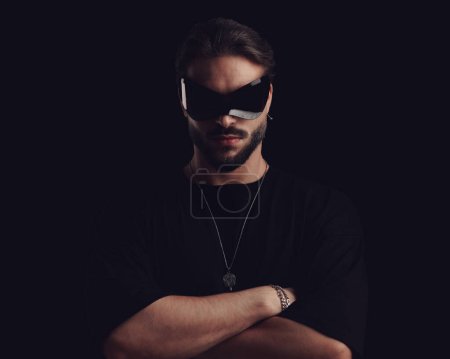 Photo for Sexy bearded man in black t-shirt crossing arms and looking forward while wearing cool sunglasses in front of black background - Royalty Free Image