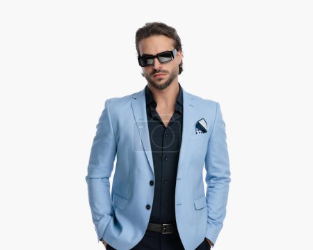 Téléchargez les photos : Portrait d'un homme d'affaires élégant avec des lunettes de soleil tenant la main dans les poches et posant devant un fond blanc - en image libre de droit