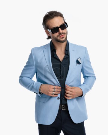 Téléchargez les photos : Cool fashion guy avec des lunettes de soleil regardant loin tout en boutonnant costume en face de fond blanc - en image libre de droit