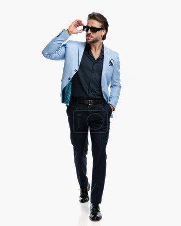 Photo for Attractive elegant man in suit with open collar shirt arranging sunglasses, looking to side and walking with hand in pocket - Royalty Free Image