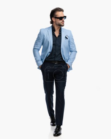 Téléchargez les photos : Homme d'affaires sexy en costume avec chemise déliée marchant avec les mains dans les poches et regardant vers le côté en face de fond blanc en studio - en image libre de droit