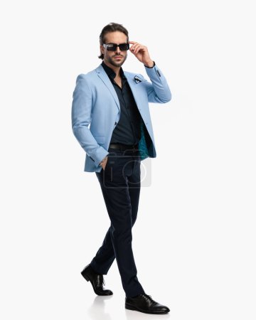 Téléchargez les photos : Homme de mode avec chemise à col ouvert marche avec la main dans la poche et la fixation des lunettes de soleil en face de fond blanc en studio - en image libre de droit