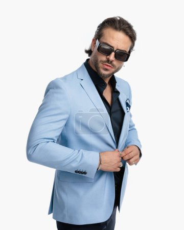 Foto de Retrato de hombre barbudo elegante con gafas de sol ajustando traje azul y mirando hacia adelante en frente de fondo blanco en el estudio - Imagen libre de derechos