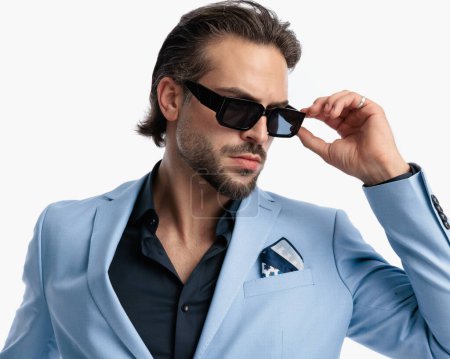Téléchargez les photos : Élégant homme barbu avec chemise à col ouvert regardant vers le côté tout en ajustant les lunettes de soleil en face de fond blanc - en image libre de droit