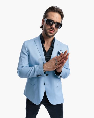 Téléchargez les photos : Sexy élégant homme d'affaires avec des lunettes de soleil frottant les paumes et regardant vers le côté en face de fond blanc - en image libre de droit