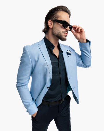 Téléchargez les photos : Sexy jeune homme d'affaires avec la main dans la poche regardant vers le côté et la fixation des lunettes de soleil en face de fond blanc - en image libre de droit