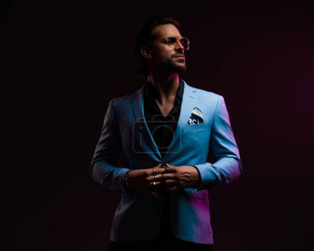 Téléchargez les photos : Homme d'affaires confiant avec des lunettes regardant vers le côté et boutonnage costume en face de fond noir avec des lumières violettes - en image libre de droit