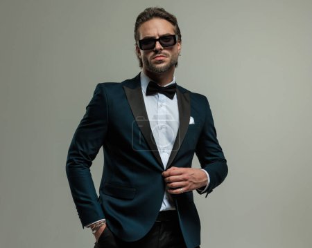 Photo for Elegant young man in tuxedo with sunglasses holding hand in pocket and unbuttoning tux in front of grey background - Royalty Free Image