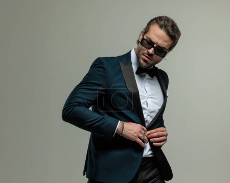Photo for Unshaved young man with sunglasses looking to side and adjusting tuxedo while posing in front of grey background - Royalty Free Image