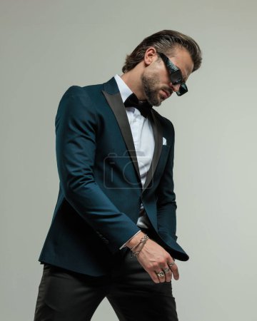 Photo for Handsome fashion man with sunglasses holding hand in pocket, looking down and posing in a cool way on grey background - Royalty Free Image