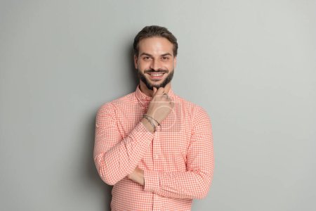 Téléchargez les photos : Jeune homme heureux réfléchi pliant les bras, touchant la barbe et pensant tout en souriant devant fond gris - en image libre de droit